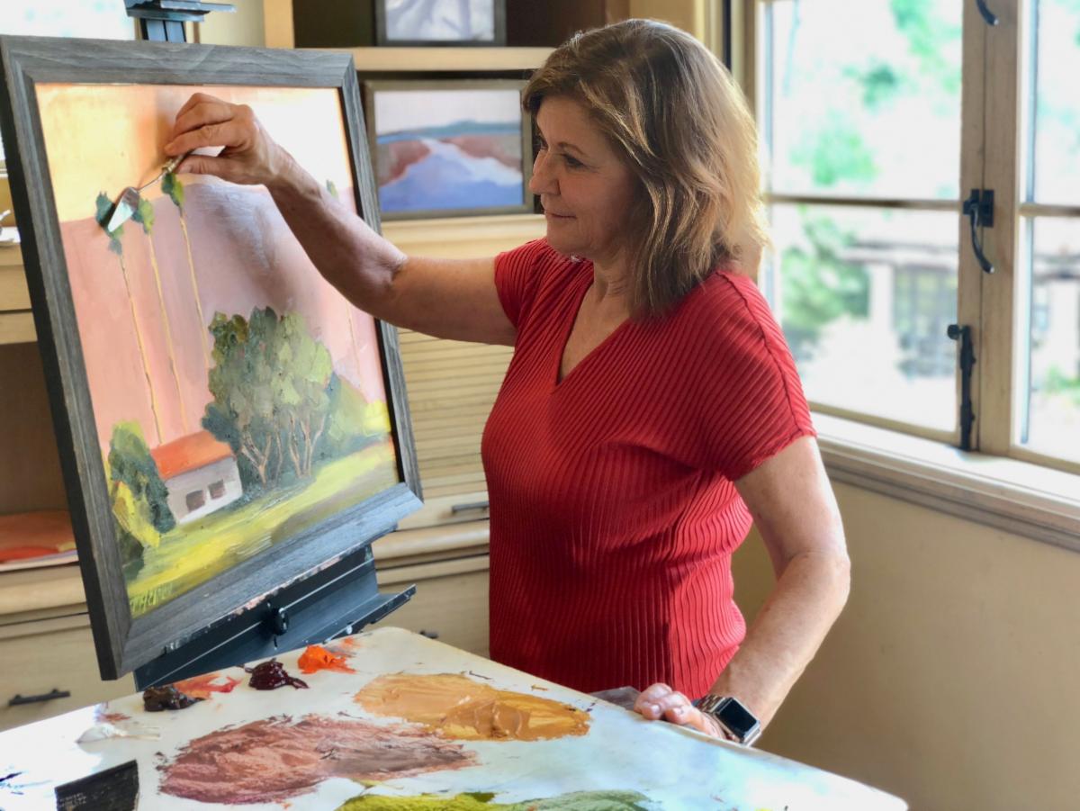 Woman standing painting a landscape on a canvas.