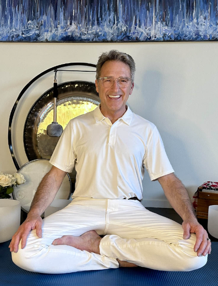 Man dressed in white shirt and white pants seated on the floor with a gong behind him.