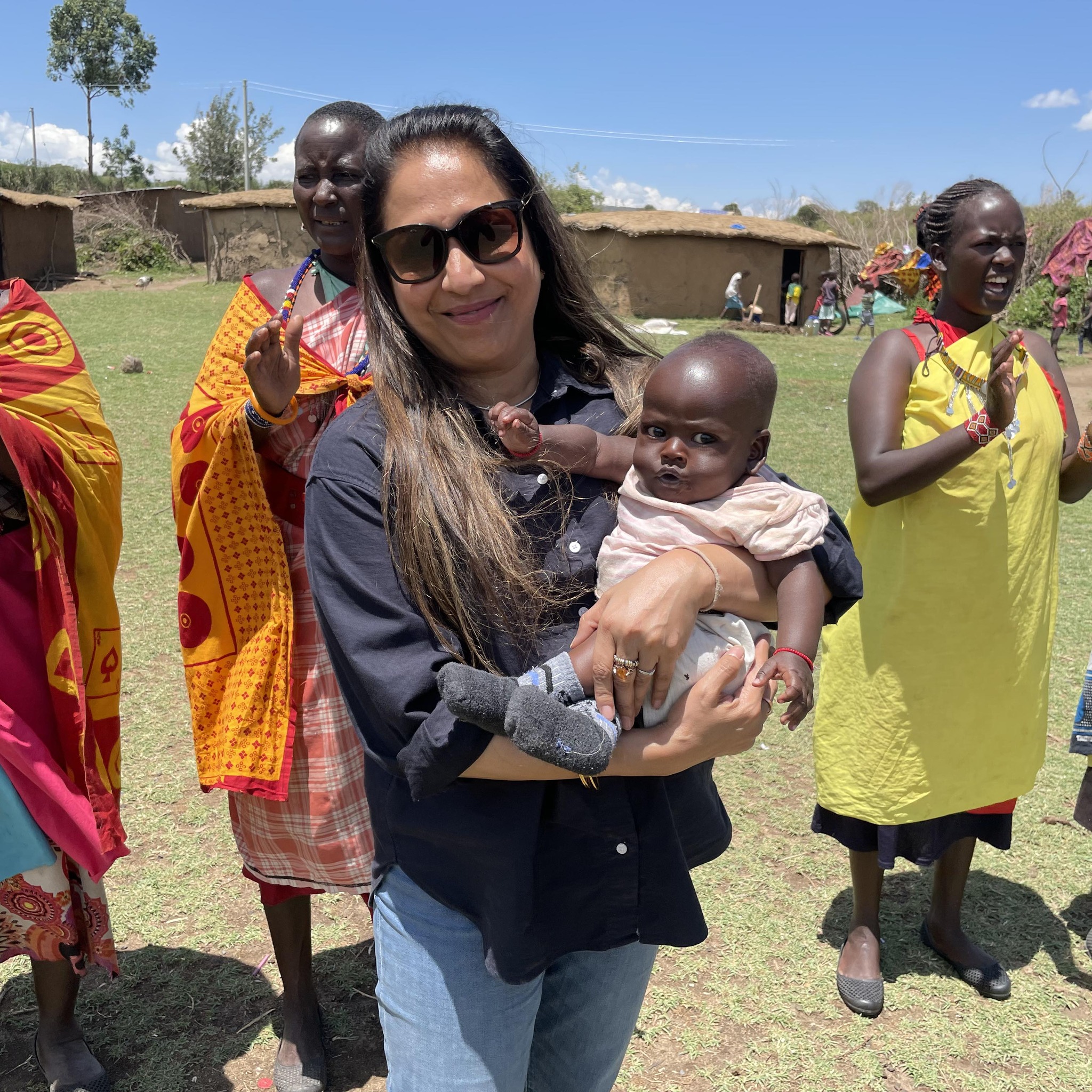 Woman holding baby outside with women in bright dresses behind her.