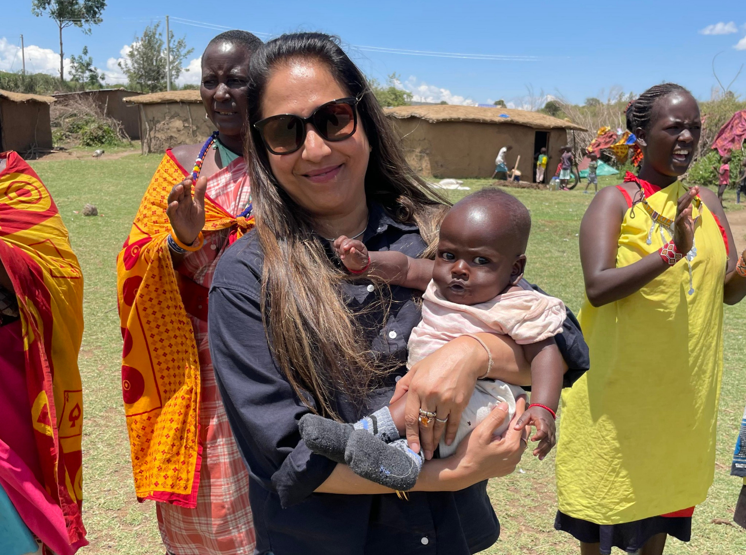 Woman holding baby outside with women in bright dresses behind her.